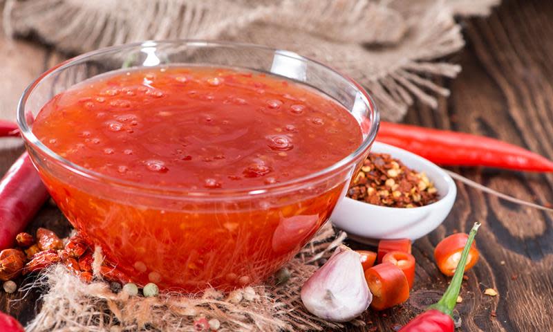 Hjemmelaget sweet chili saus - uten sukker, i gjennomsiktig skål, på mørk trebakgrunn. Omringet av chilikrydder, fersk chili og hvitløksbåter.