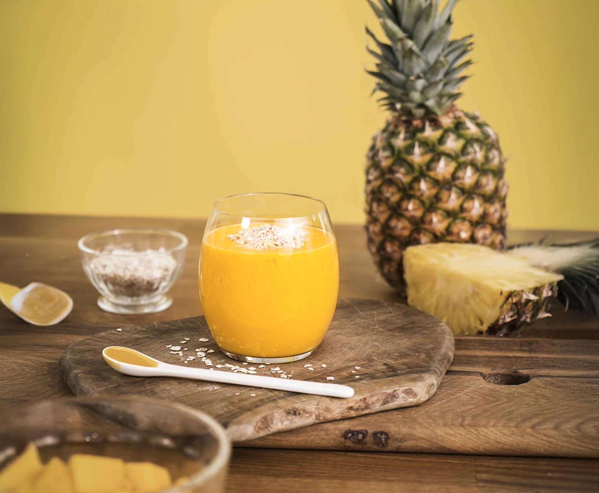 Solenergi -smoothie, gul smoothie med mango, ananas, gurkemeie og mandelmelk, i lite glass på trefjøl med fersk frukt rundt.