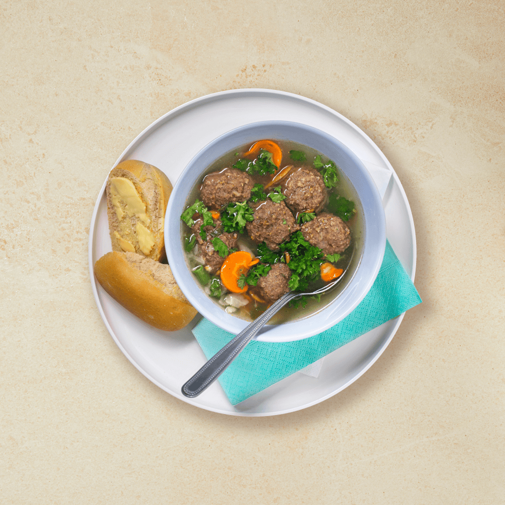 Grønnsakssuppe med kjøttboller i en hvit skål som står på en hvit tallerken med brød og servietter