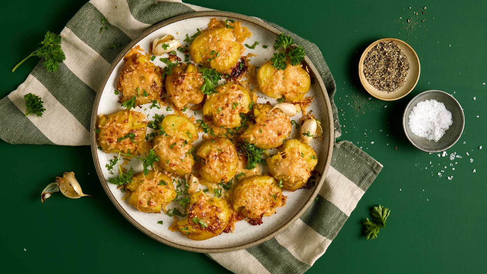 Knuste poteter med hvitløkssmør og parmesan servert på rundt lyst fat med friske urter på toppen. Små skåler salt og pepper på siden.