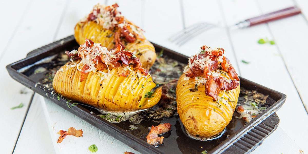 Hasselbackpoteter med bacon, servert på svart støpejernsfat med parmesan og ferske urter. Fatet står på hvitt trebord, og det er en gaffel i bakgrunnen.