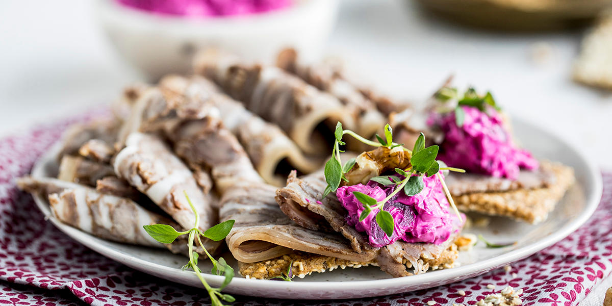 Knekkebrød med skiver av julesylte og rødbetesalat, servert på tallerken på lillamønstret serviett. Friske grønne urter på toppen.