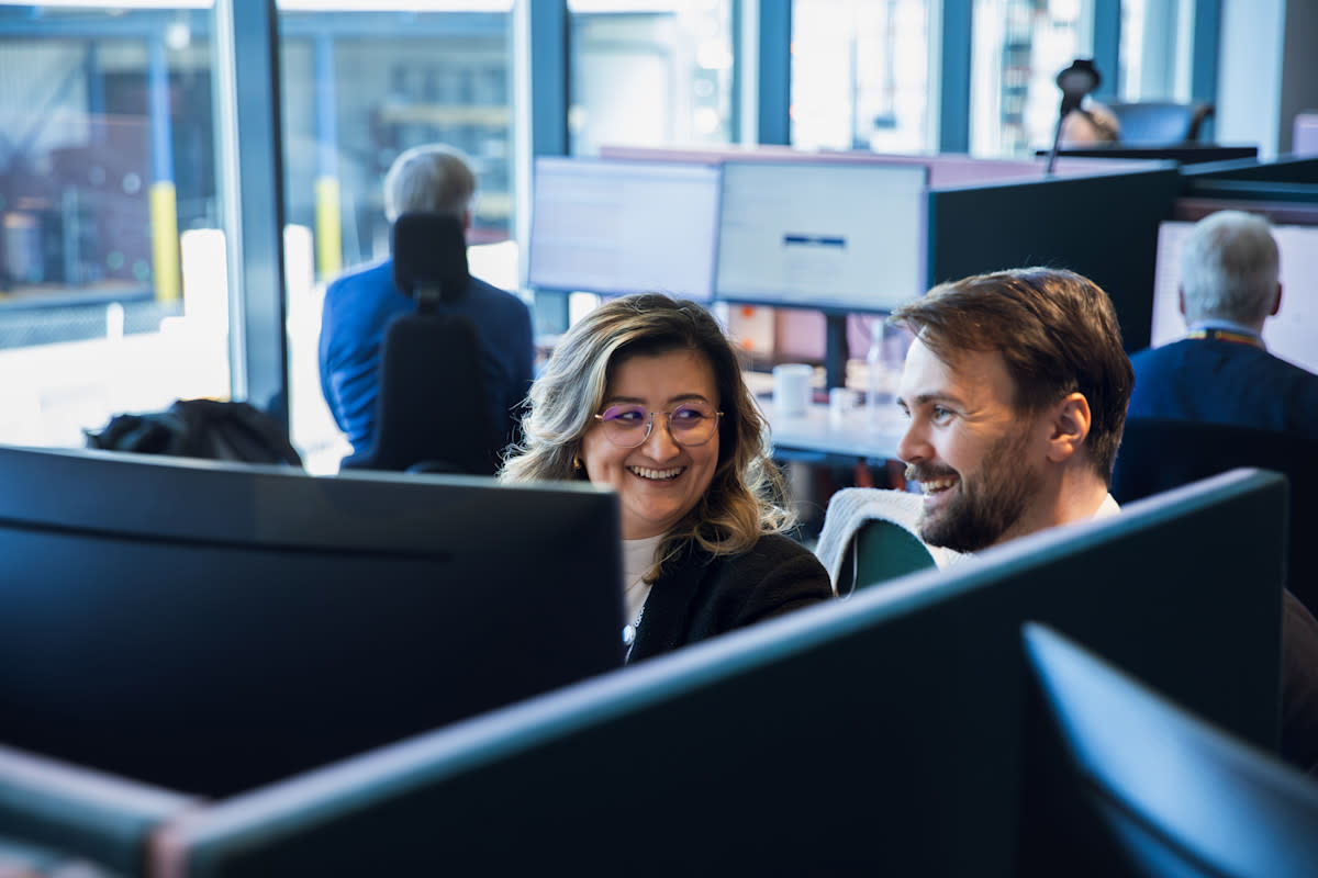 en mann og en kvinner sitter et et åpent kontorlandskap,  de smiler og ser på en PC skjerm