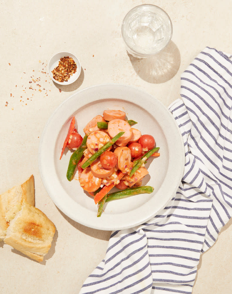 Enkel pølsegryte med paprika servert på hvit tallerken med et glass vann og lys baguette ved siden av.