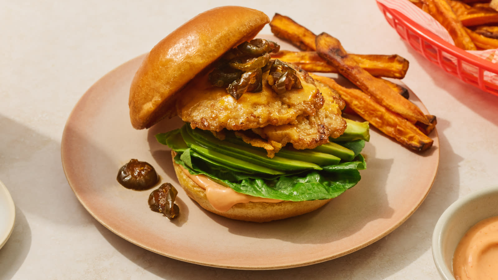 Smashed kyllingburger med søtpotetfries servert med avokado, salat, cowboy candy og srirachamayo på en rosa tallerken.