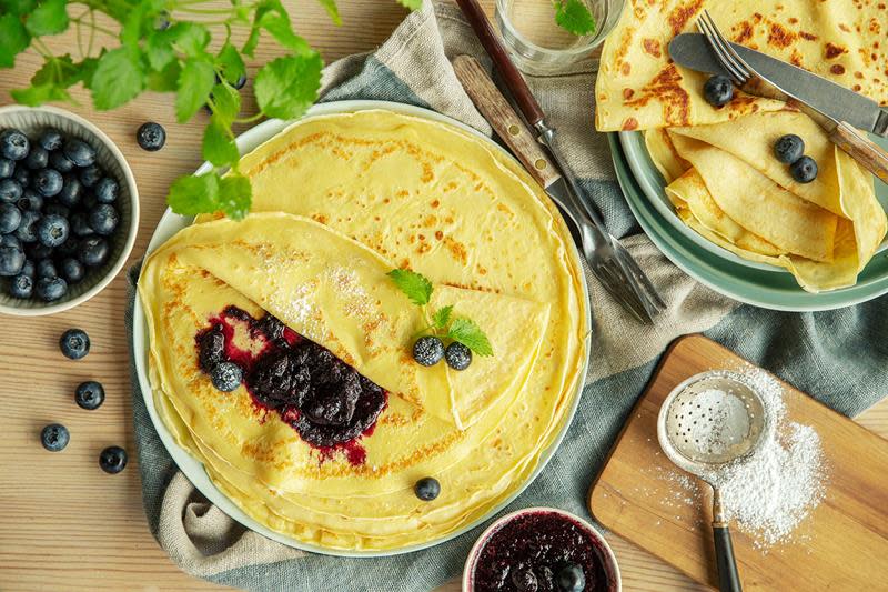 Nystekte gyldne pannekaker på lyst fat, servert med blåbærsyltetøy, ferske blåbær og et dryss melis.