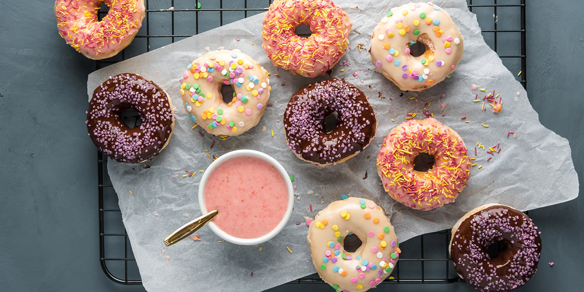 Bakte donuts med 3 typer glasur i jordbær, sjokolade og vanilje. Donutsene er dyppet i glasur, pyntet med forskjellige typer kakestrø og servert på kjølerist med hvitt bakepapir. Liten skål med jordbærglasur på siden.