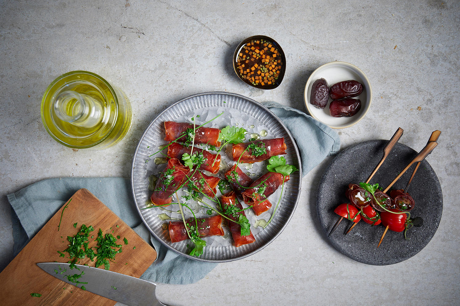 Dadler med sprø spekeskinke og hjemmelaget dip servert på rund tallerken med små skåler dipsaus og dadler rundt. Liten skjærefjøl med friske urter ved siden av.