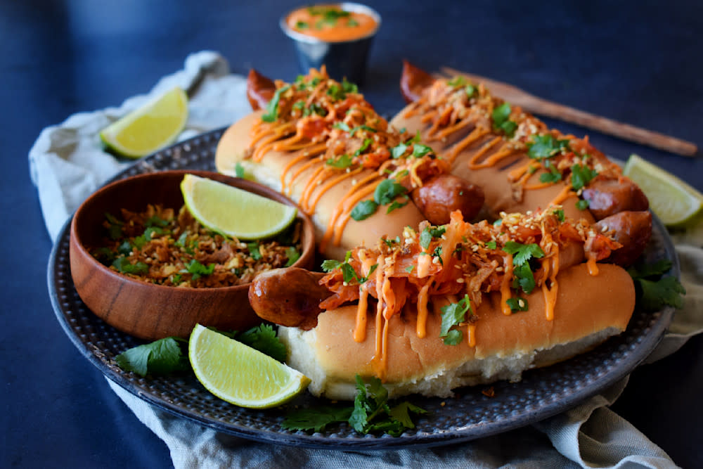 Beefy sausage med hjemmelaget kimchi og honning- chilimayo servert med crunchy topping i pølsebrød på mørk tallerken. Liten skål med chilimayo i bakgrunnen, og limebåter rundt.