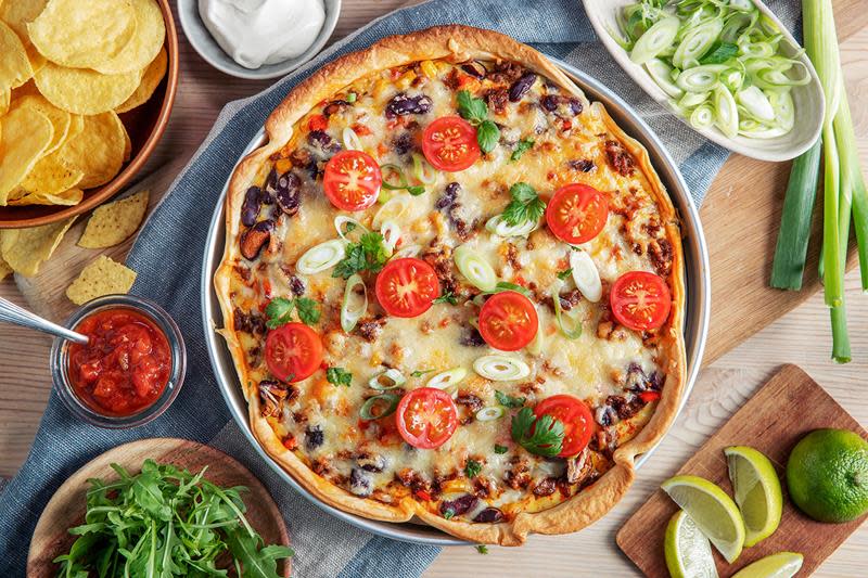 Rund tacopai med kjøttdeig toppet med ost, ferske tomater og snittet vårløk. Paien står på middagsbord omringet av skåler med salsa, ferske urter, limebåter, nacho chips og rømme. Blått linhåndkle.