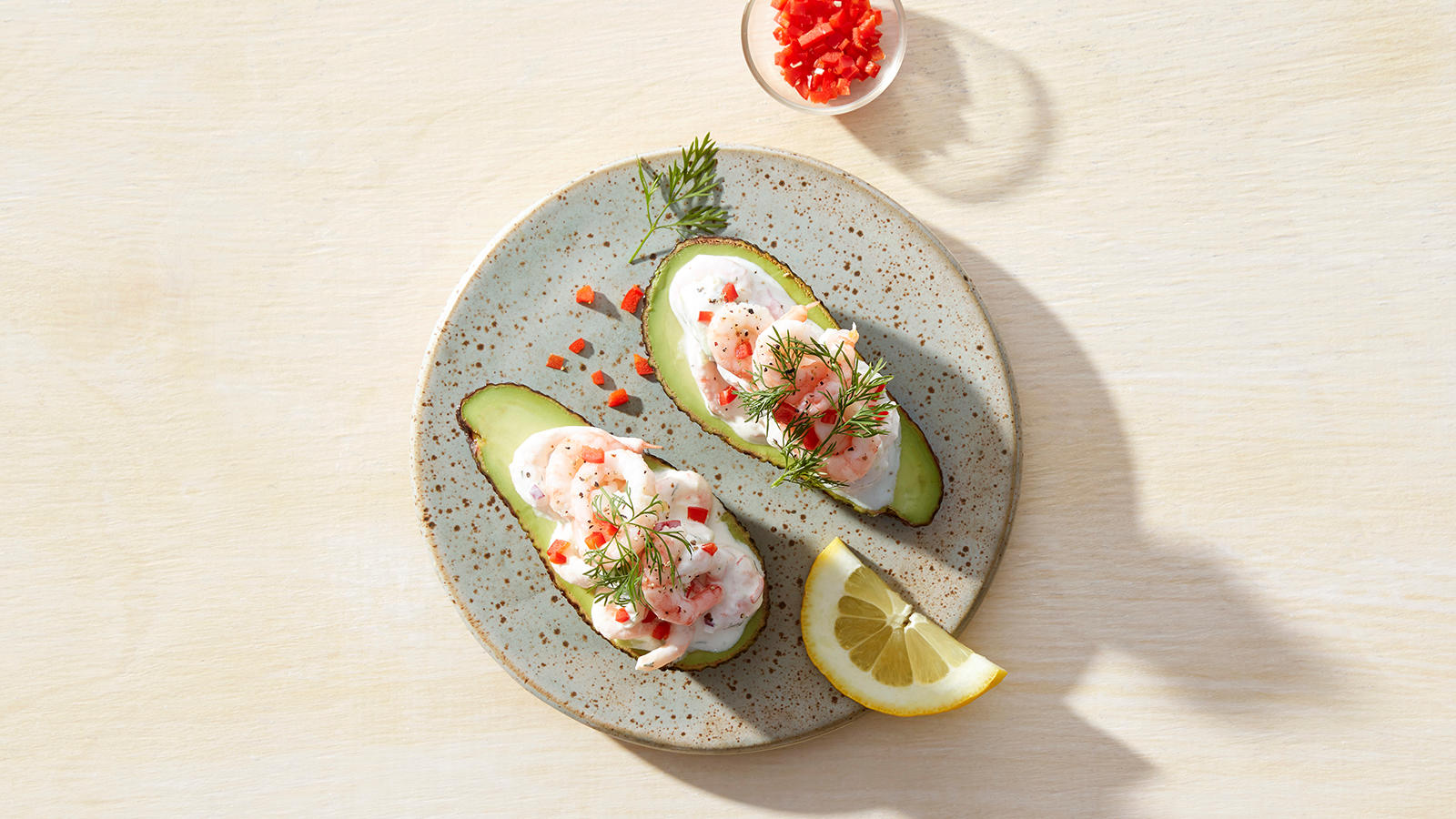 Fylt avokado med reker, servert med chili, frisk dill og en halv sitronskive på lyseblå keramikktallerken, på lyst trebord. Liten skål med rød chili ved siden av.