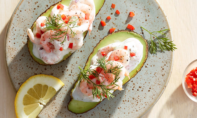 Fylt avokado med reker, servert halve med chili, dill og sitronskive på lyseblå prikkete tallerken på lyst trebord.