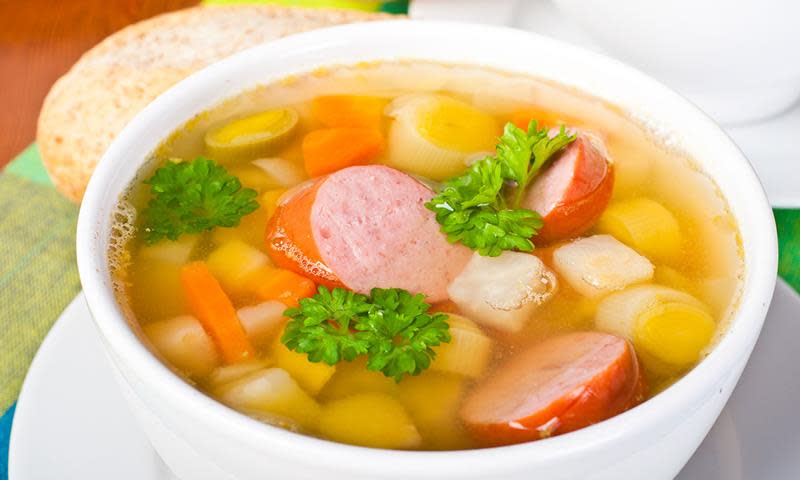 Kjøttsuppe med pølse og fargerike rotgrønnsaker, i hvit suppeskål, med baguette på siden, og kruspersille på toppen.