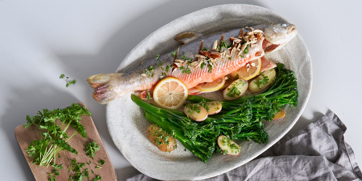 Fat med hel røye med hode og hale. Fisken er fylt med sitronskiver og dill, og den er toppet med hakkede mandler. Servert med halve poteter med urter og brokkolini.