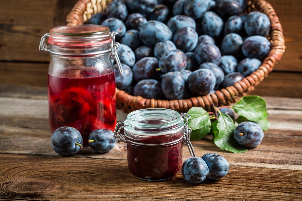 Hermetiserte plommer på glass, i rød lake står på rustikk trebord. Kurv med blå plommer i bakgrunnen, og glass med plommesyltetøy ved siden av.