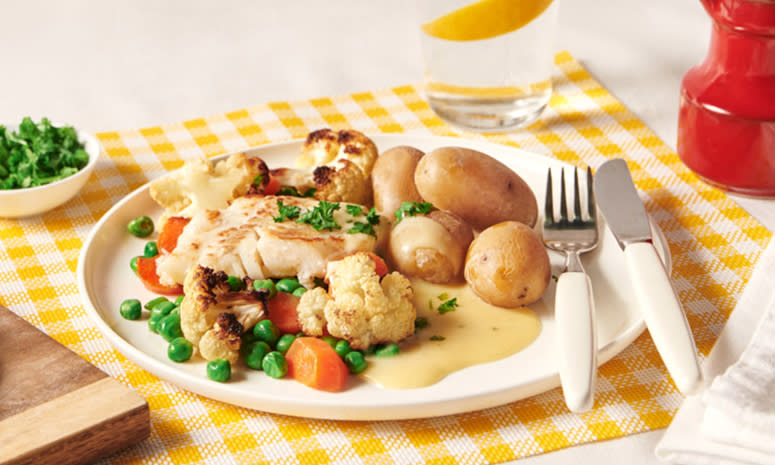 Lettsaltet torsk med sandefjordsmør, poteter, erter og gulrot, servert på en hvit tallerken med bestikk som ligger på en gul og hvit rutete duk.