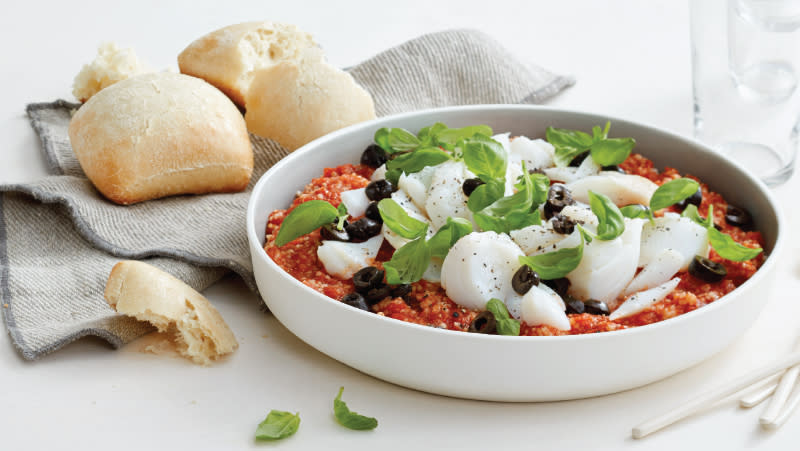 Marrokansk torskegryte med couscous, tomatsaus og oliven servert i hvit form med fersk basilikum på toppen, og små ciabatta ved siden av på en linserviett.