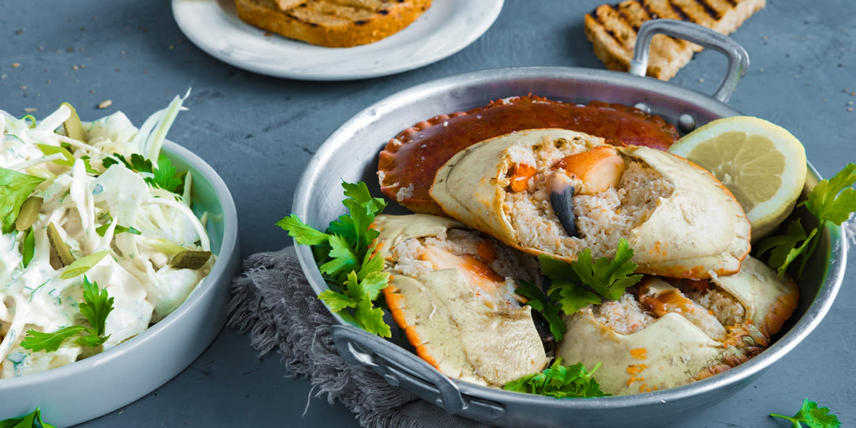 Krabbeskjell med klo servert i metallkjele med bladpersille og sitronskive. Skål med hjemmelaget potetsalat ved siden av, og tallerken med ristet brød i bakgrunnen.