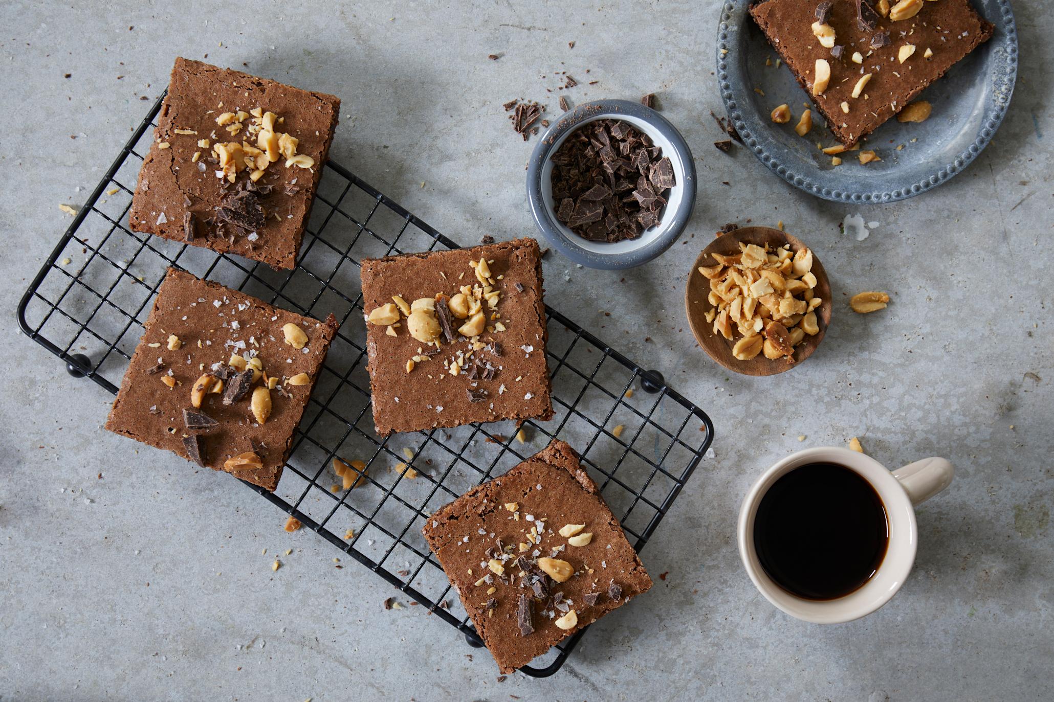 Glutenfri brownies med kaffe servert i ruter på svart kjølerist, og pyntet med hakkede peanøtter og mørk sjokolade. En lys kopp med mørk kaffe ved siden av.