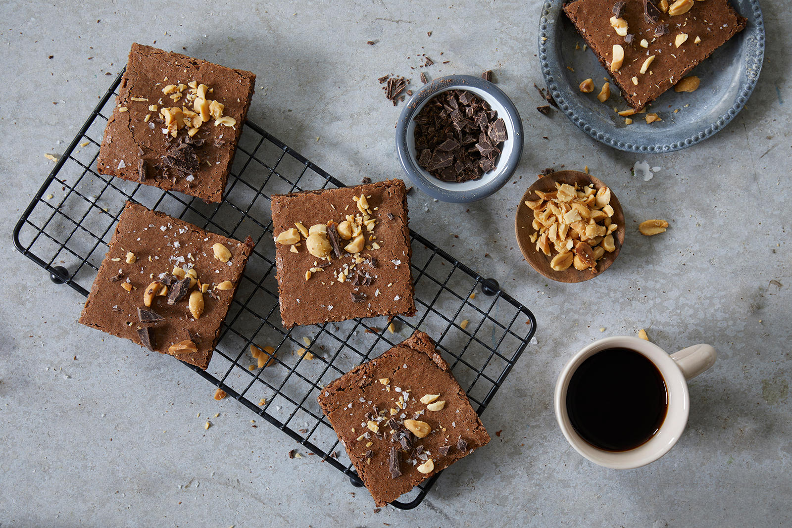 Glutenfri brownies med kaffe servert i ruter på svart kjølerist, og pyntet med hakkede peanøtter og mørk sjokolade. En lys kopp med mørk kaffe ved siden av.