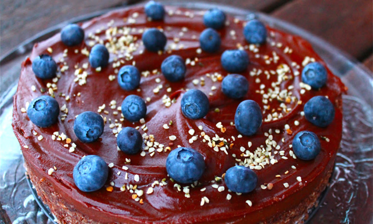 Raw sjokoladekake med dadler og nøtter pyntet med friske blåbær og hempfrø, og servert på kakefat i glass.