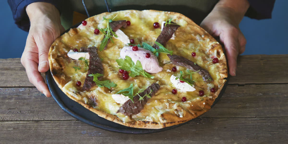 Hvit pizza med skiver av reinsdyrfilet, snøfrisk, pisket tyttebærrømme, ruccola og ferske tyttebær servert på mørk tallerken holdt frem av to hender.