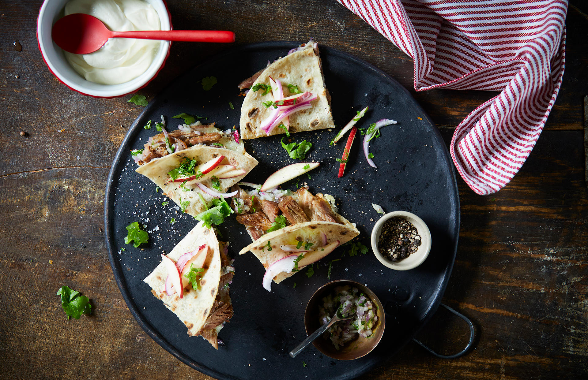 Quesadilla med pinnekjøtt, persillesalsa og eplesalat delt i fire og servert på svart tallerken med ferske urter, epleskiver og skivet rødløk på toppen. Små skåler med persillesalsa, rømme og nykvernet pepper ved siden av.