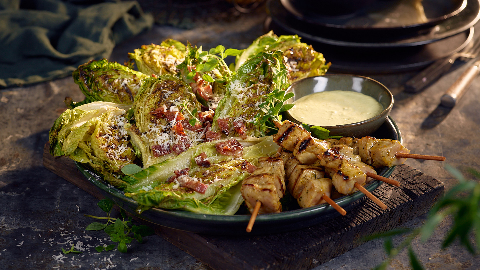 Grillet cæsarsalat med hjertesalat, parmesan og bacon servert med kyllingspyd på mørk tallerken. Liten skål dressing på siden.