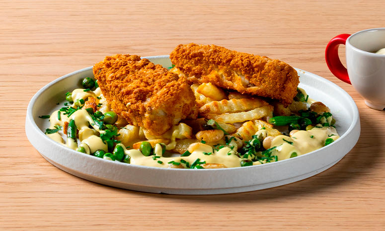 Fish and chips med hollandaise og bønnemiks servert på hvit tallerken på lyst trebord. Liten kopp med rødt håndtak ved siden av.