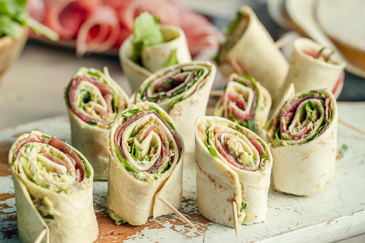 Lefserull med spekeskinke og avokadokrem, skjært i skiver og servert på lys rustikk trefjøl. Fat med spekeskinke i bakgrunnen.