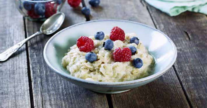 Havregrøt med Coop Gullbygg servert i keramikkskål med friske bringebær og blåbær. Skålen står på mørkt trebord med skje ved siden av og et glass friske bær i bakgrunnen.