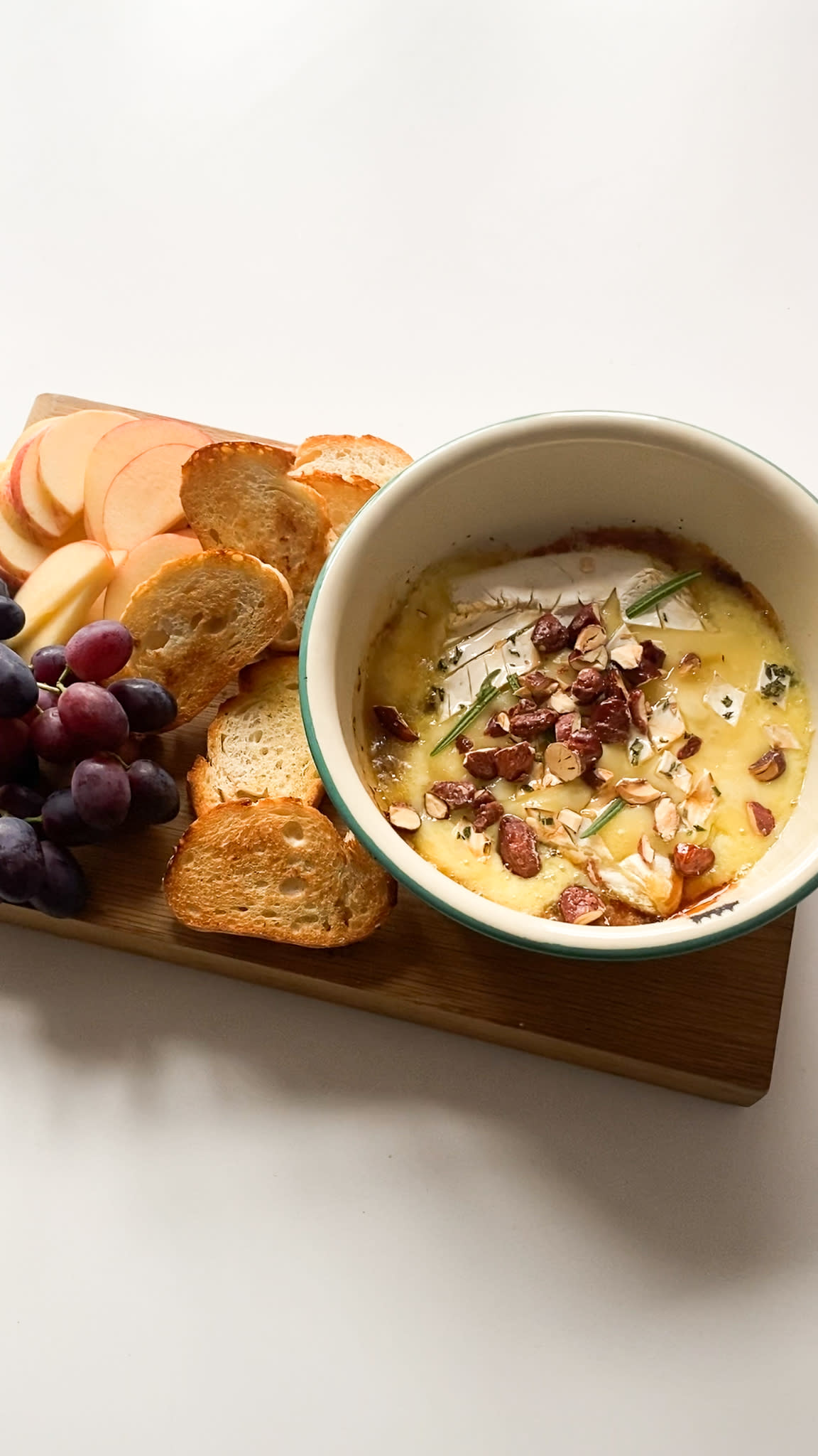 Bakt brie med karamelliserte mandler, servert i rund ildfast form på trefjøl med ristet baguetteskiver, røde druer og epleskiver ved siden av.