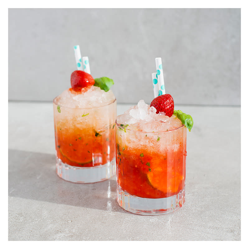 To glass med knust is, rød drikke og limebåter pyntet med et basilikumblad, et jordbær og to blå og hvite sugerør.