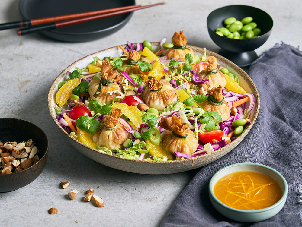 Asiatisk salat med dumplings og appelsinsaus servert i lav lys keramikkbolle, med små skåler mandler, edamame og appelsinsaus på siden.