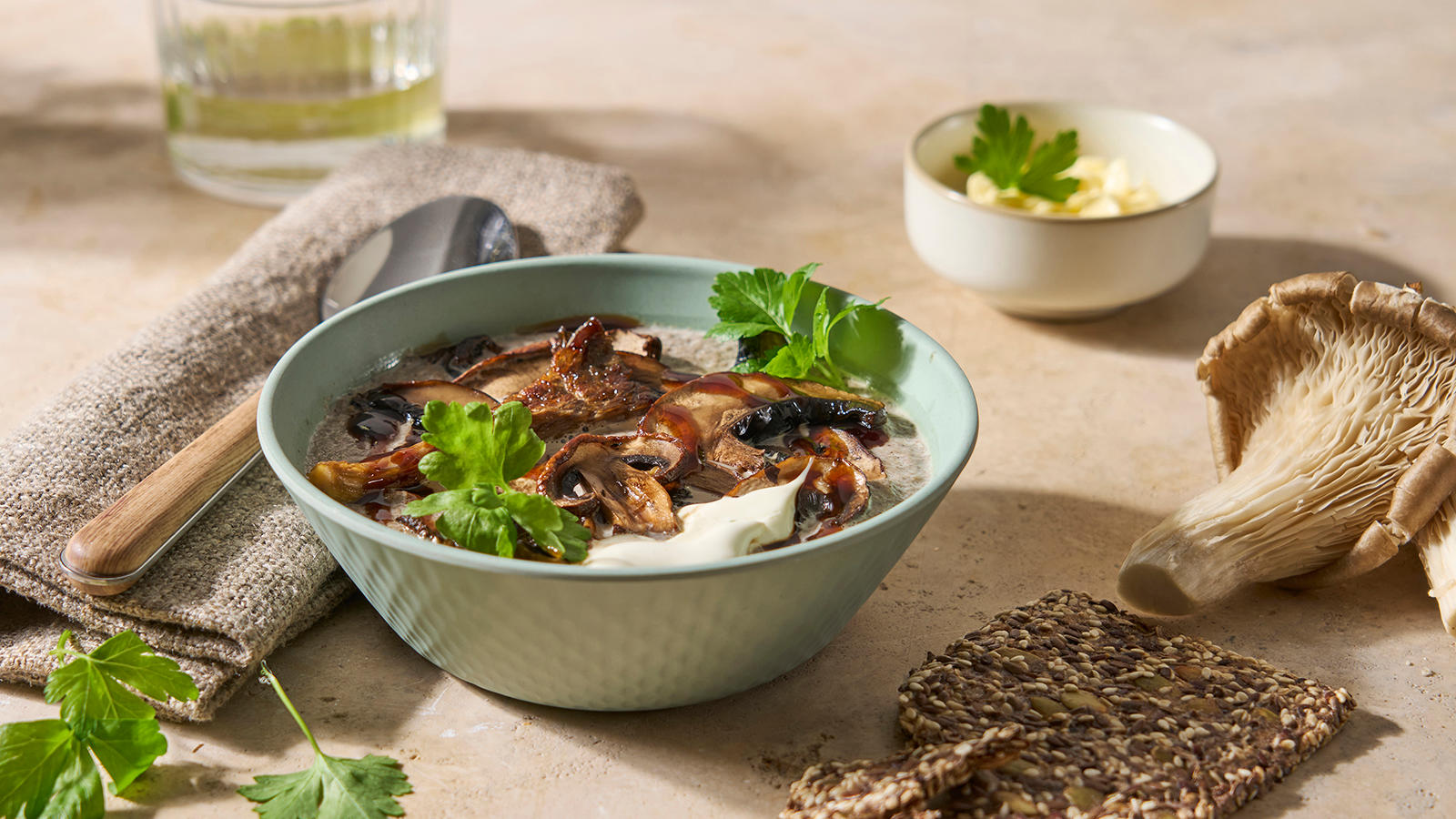 Kremet soppsuppe med sjampinjong, østerssopp og portobellosopp servert i mintgrønn suppeskål med frisk bladpersille. Frøknekkebrød, friske urter og østerssopp rundt.