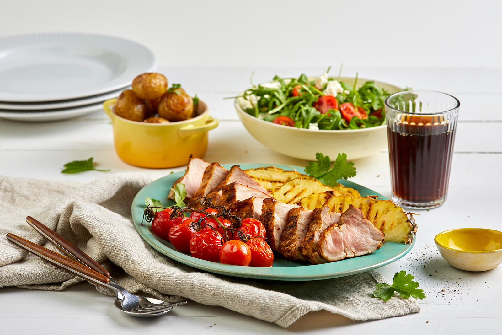 Ytrefilet av svin med ovnsbakte småpoteter, grillet ananas og salat med fetaost, servert på turkis asjett med grillede cherrytomater. Skåler med sommersalat og ovnsbakte poteter i bakgrunnen.