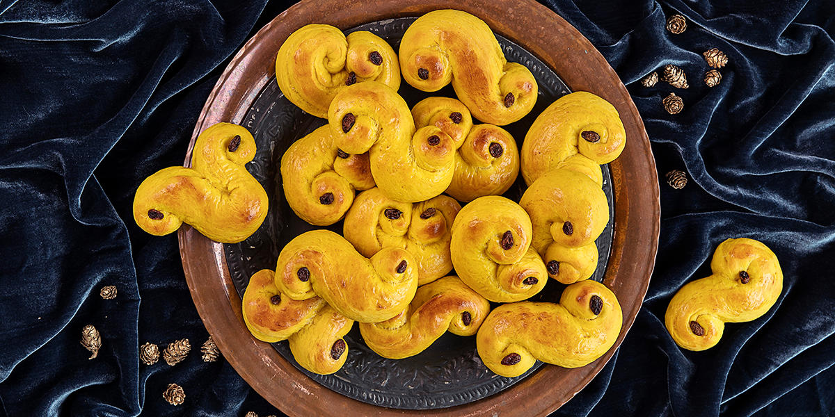 Luftige lussekatter i forskjellige former, servert på mørkt jernfat på mørkeblå fløyelsduk med små kongler strødd rundt.