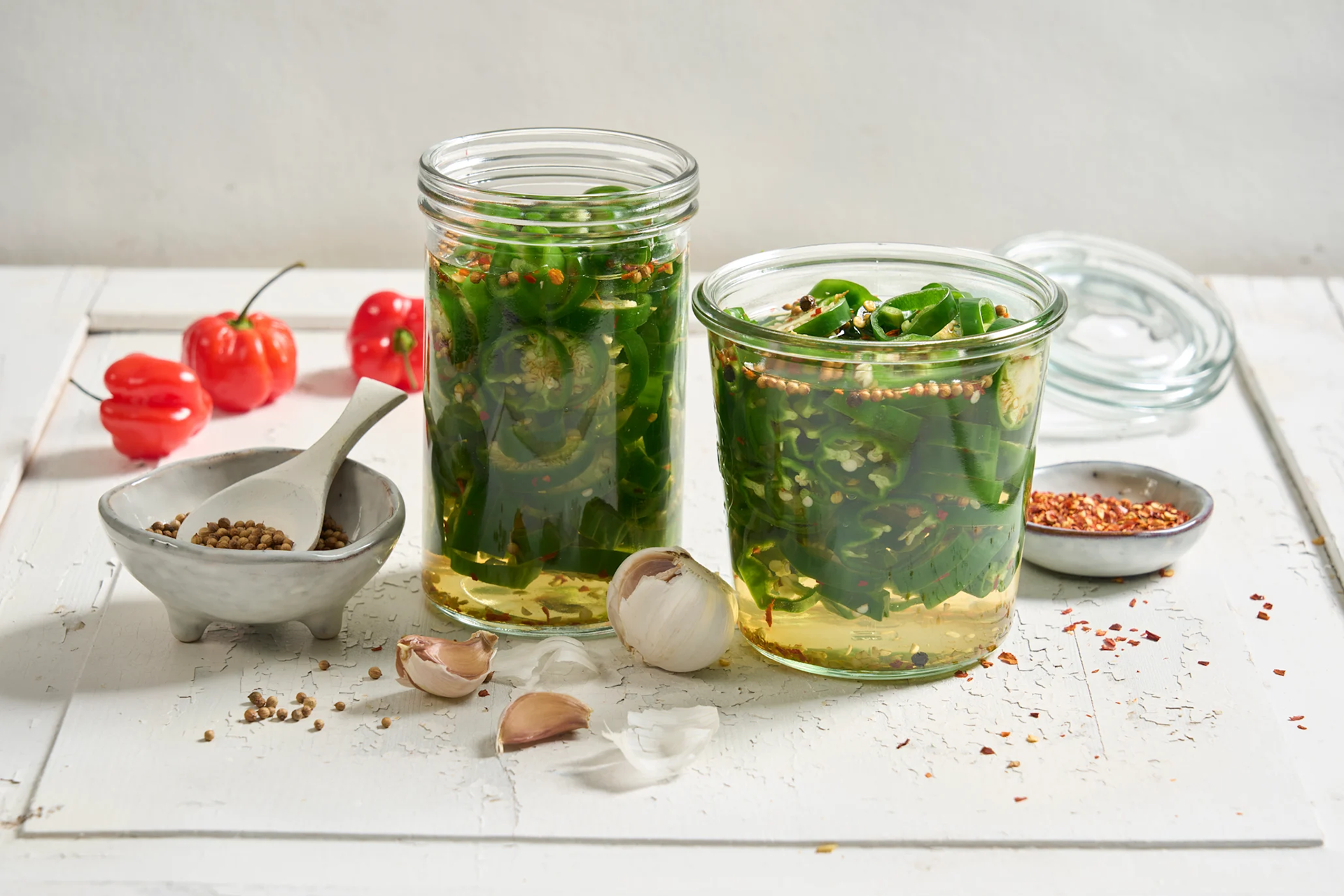 Sylta padron med korianderfrø og chili, skåret i skiver og lagt på to forskjellige sylteglass. Hele røde chili, hvitløk og små skåler med krydder ved siden av.