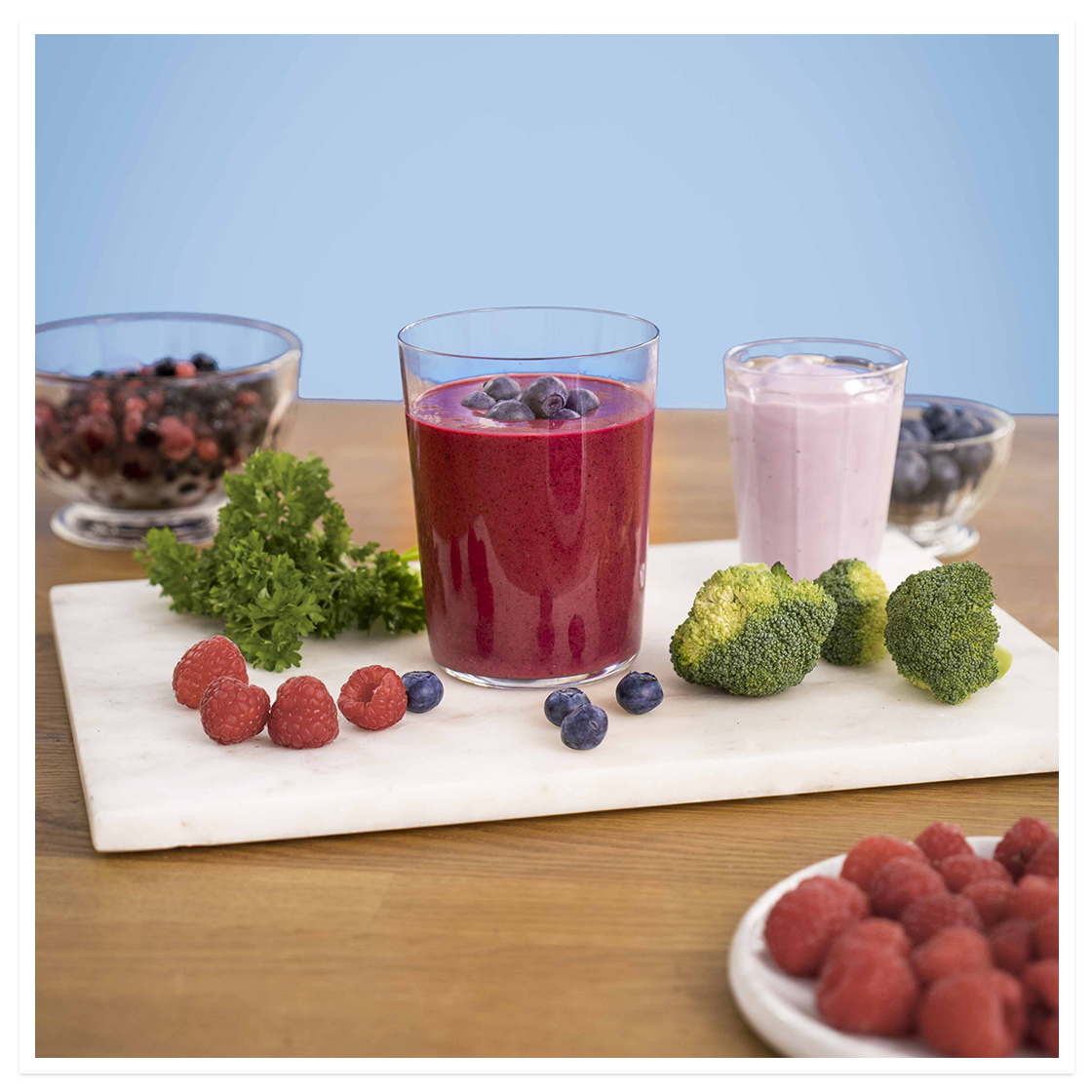 Blåmandag - smoothie, lilla smoothie med skogsbærblanding, blåbær, brokkoli og persille, står på hvit fjøl på frokostbord. Ferske bær og grønnsaker rundt.