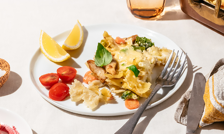 kylling, pasta og brokkoli dekket av smeltet ost på hvit tallerken sammen med tomater, sitronbåter og en gaffel.