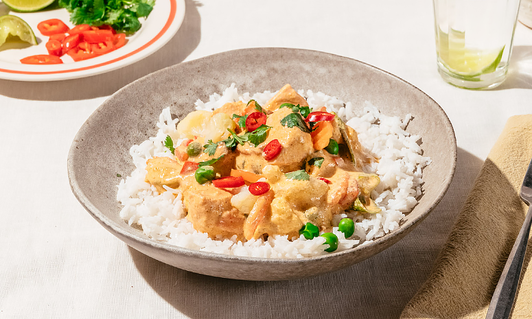 Laks i red curry-saus med ris server i en grå skål med et glass vann og en tallerken i bakgrunnen med oppskjært chili og limebåter, på høyre side av skålen ligger bestikk på et beige tørkle