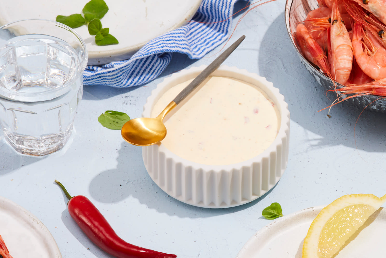 Chiliaoli til reker servert i hvit marmorskål med riller, og en liten skje i grått og gull på toppen. Skålen står på et dekket middagsbord med frisk chili, sitronbåt, ferske uter og en skål ferske reker.