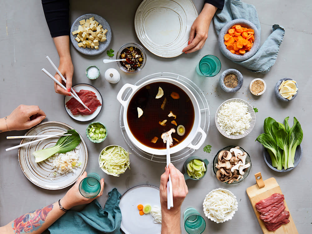 Shabu shabu – Japansk fondue, servert i hvit kjele midt på bordet med en rekke tilbehør og tallerkner rundt, samt 3 mennesker som spiser.