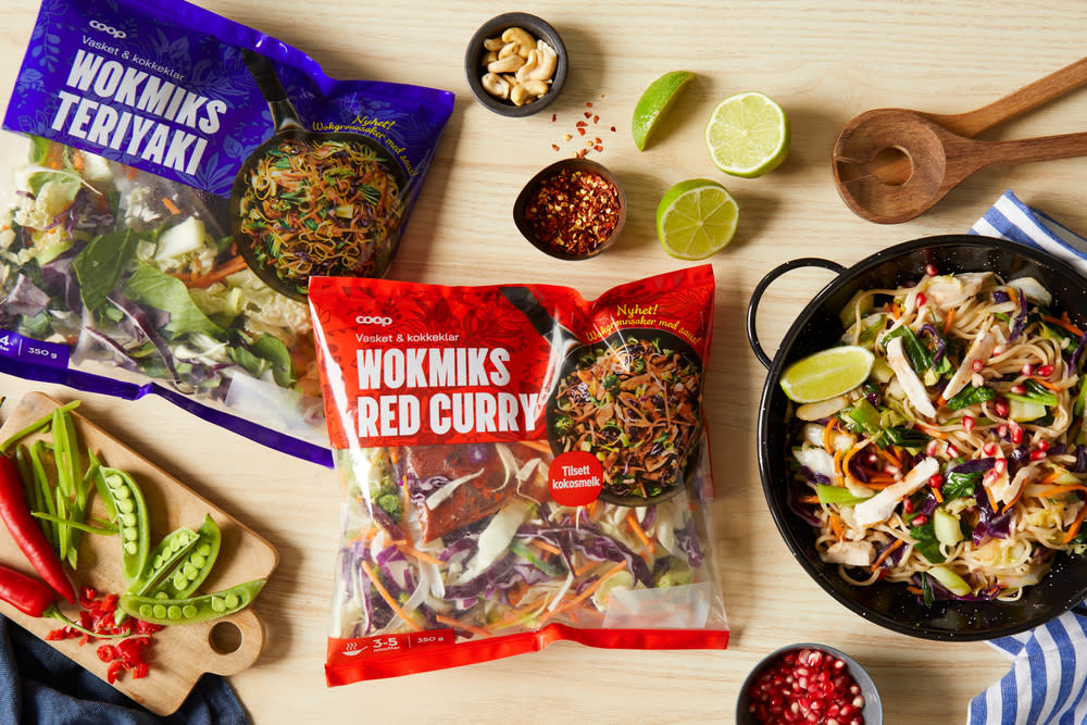 Coop wokmiks Teriyaki og Coop wokmiks Red curry i emballasje ligger på lyst trebord ved siden av en skål med tilberedt wok.
