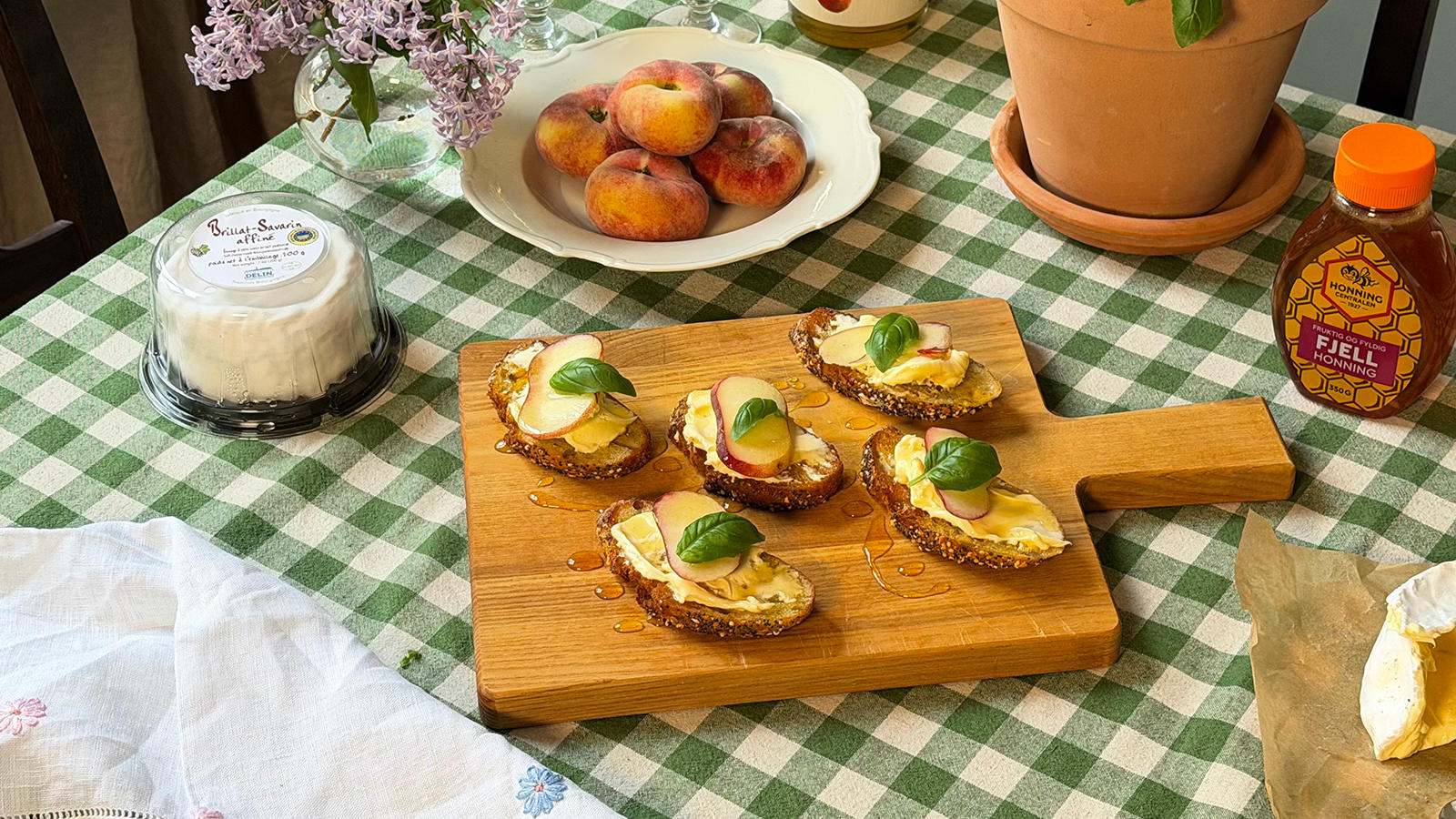 Crostinis med Brillat-Savarin og hvite ferskner i skiver, frisk basilikum og honning servert på firkantet trefjøl på grønnrutet duk.