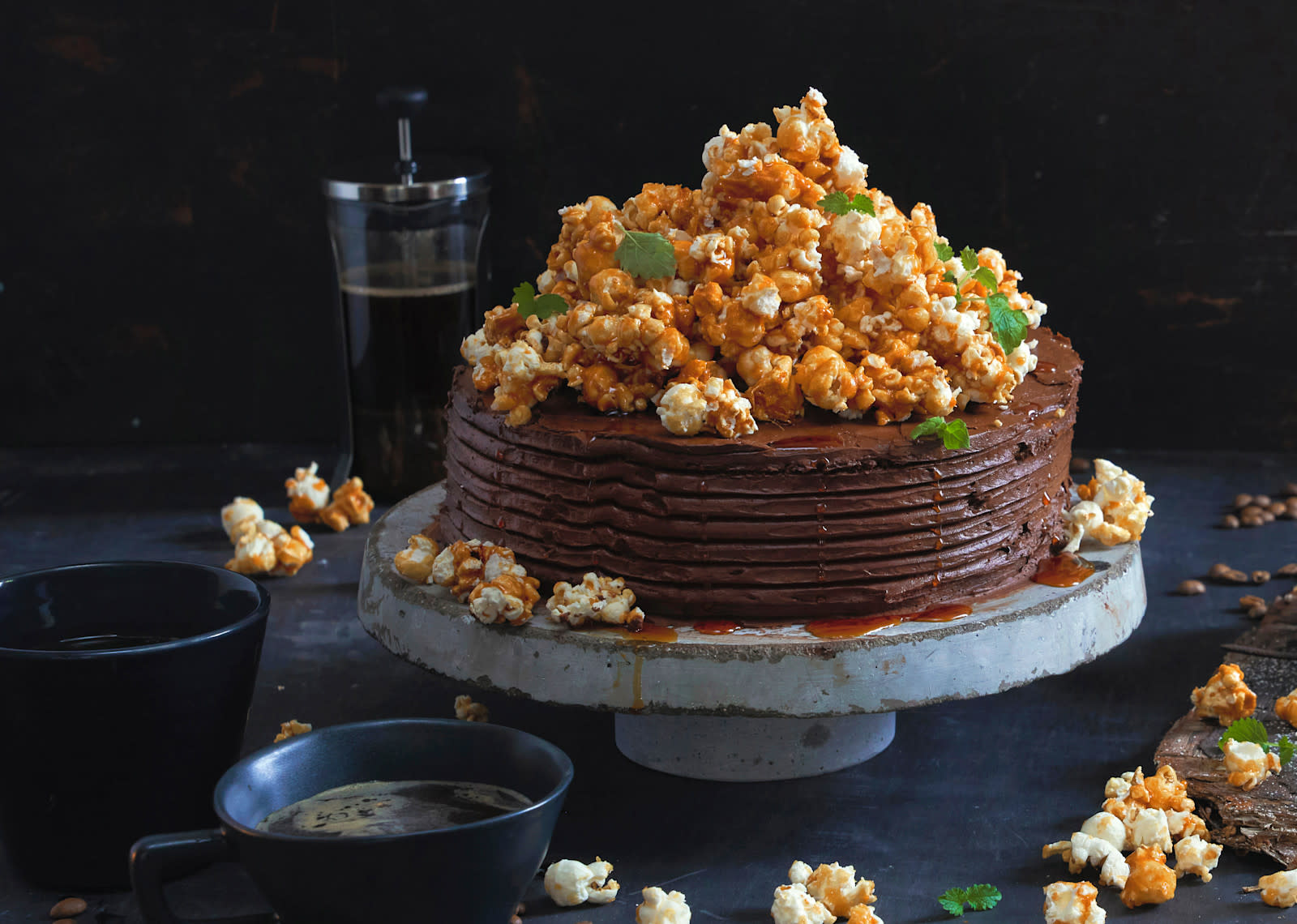 Sjokoladekake med mørk sjokoladekrem og karamellpopcorn servert på stettfat i metall, med karamellsaus ringlet over. Kopper med kaffe ved siden av.