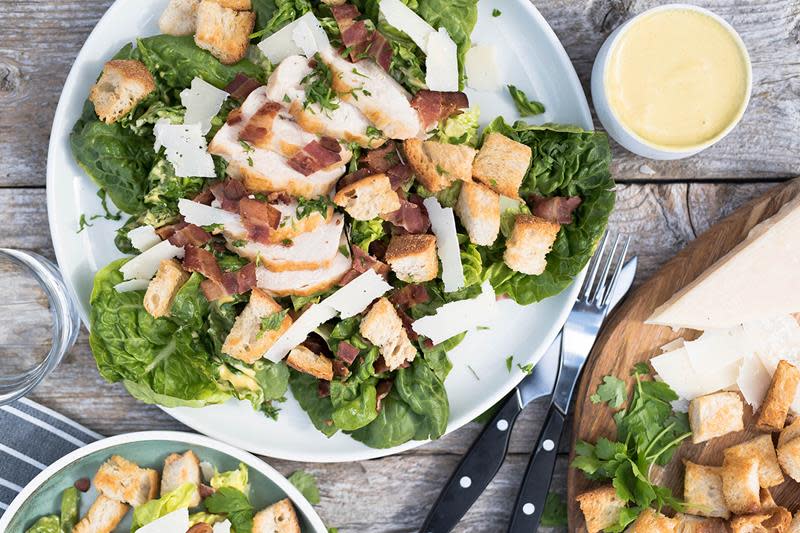 Cæsarsalat med kyllingbryst, parmesan og bacon anrettet på hvitt fat, med dressing i skål ved siden av. Rustikk trebakgrunn.