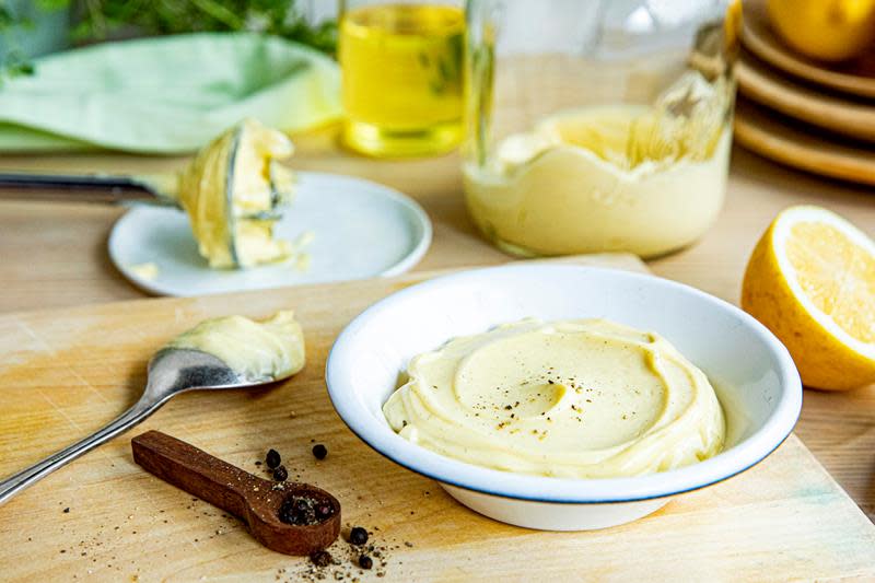 Hjemmelaget majones pyntet med nykvernet pepper i lys liten serveringsskål, på trefjøl. Flaske med olje, fersk sitron, og hel pepper ved siden av.