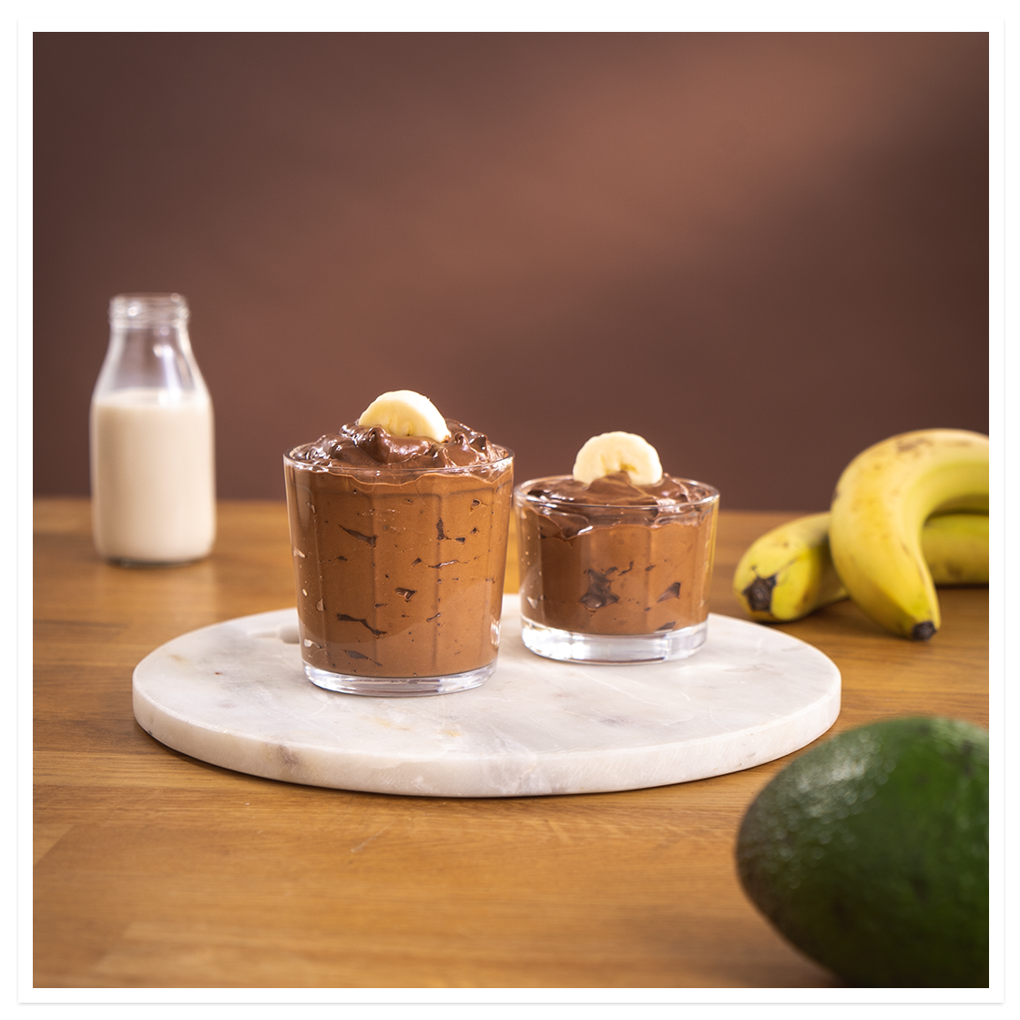 Sjokofest -smoothie med banan, avokado og mandelmelk, i to små glass, pyntet med bananskiver. Hvit marmorfjøl på trebord.