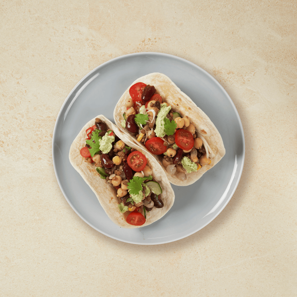 Vegetartaco i to små tortillalefser på grå tallerken. Lefsene er fylt med forskjellige typer bønner, guacamole, ferske tomater og koriander.